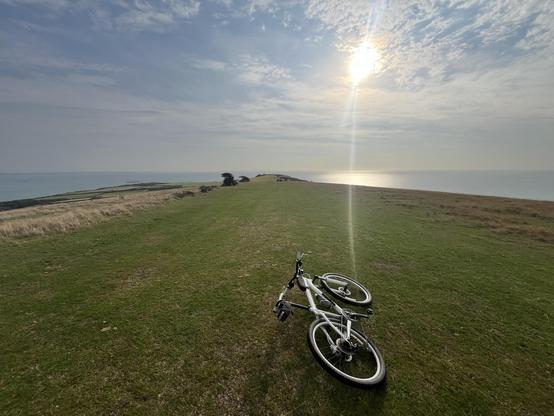 A bicycle lies on a grassy hill overlooking a calm sea under a partly cloudy sky. The sun is visible, shining brightly, and a ship can be seen in the distance along the water. The scene conveys a serene outdoor environment.