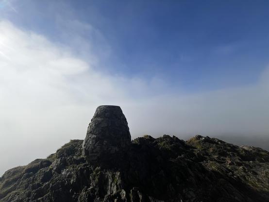 A stone summit marker sits atop a rocky outcrop, surrounded by a misty landscape under a bright blue sky.