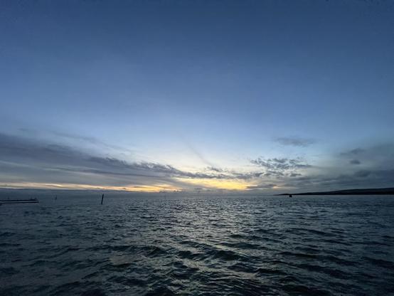 A serene view of the ocean at dusk, with a gradient sky transitioning from blue to warm hues near the horizon. Subtle waves can be seen on the water, along with distant silhouettes of structures and poles.