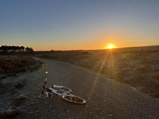A bicycle lies on a gravel path with a scenic view of a sunrise in the background. The sun is low on the horizon, casting warm light over the landscape, with trees visible in the distance.
