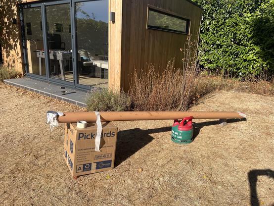 A brown pole rests on a cardboard box labeled "Pickfords" in an outdoor setting. A green gas canister is visible nearby. This is the basis of a pin hole camera