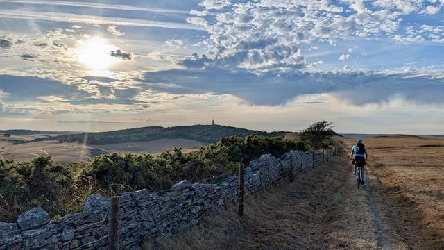 A cyclist rides along a dirt path surrounded by dry grass and shrubs, with a rocky fence on one side. The landscape features rolling hills with Hardy Monument in the distance, under a sky filled with clouds and sunlight.