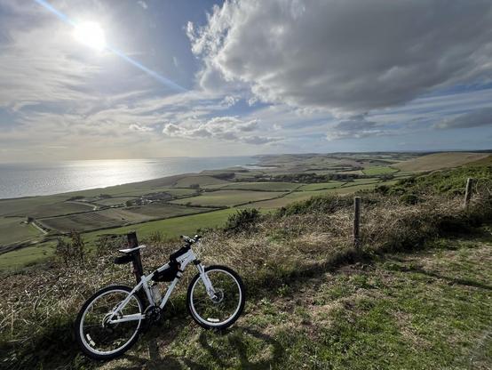 A white bicycle is parked on a grassy hill overlooking a scenic landscape with fields and a coastline under a partly cloudy sky. The sun is shining brightly, reflecting on the water.