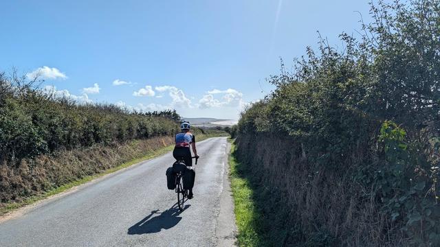 A cyclist riding on a rural road surrounded by hedges, under a blue sky with clouds.