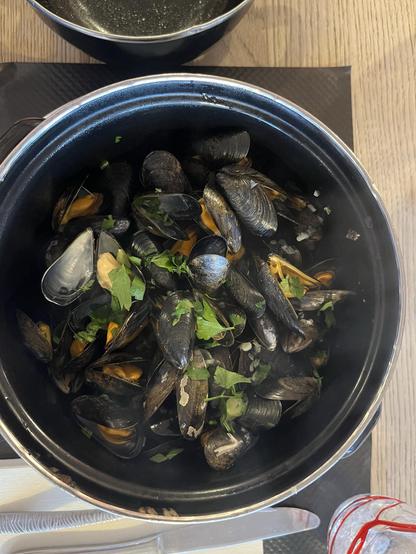 A black pot filled with cooked mussels garnished with fresh parsley sits on a wooden table. A second pot is visible in the background. There are hints of steam rising from the mussels, suggesting they are freshly prepared.