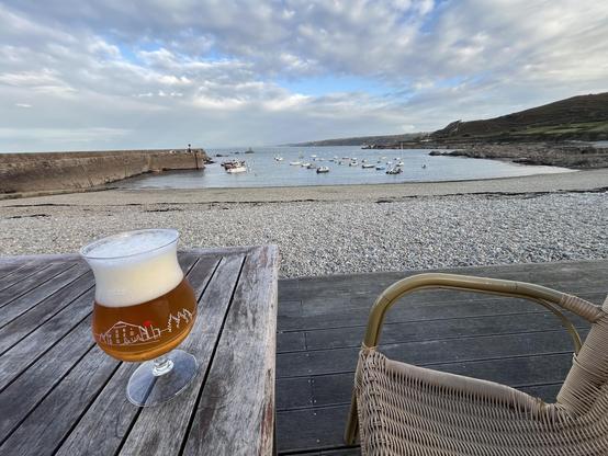 A glass of beer sits on a wooden table, overlooking a calm beach with pebbles and moored boats. The sky is partly cloudy, suggesting a tranquil seaside setting.