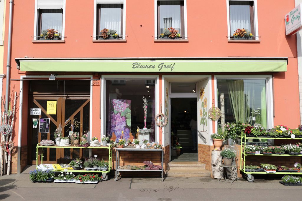 Fassade eines Blumenladens mit orangefarbener Außenwand, drei Fenstern mit Blumenkästen und einer grünen Markise, unter der bunte Pflanzen- und Blumenarrangements auf Tischen und Regalen stehen.