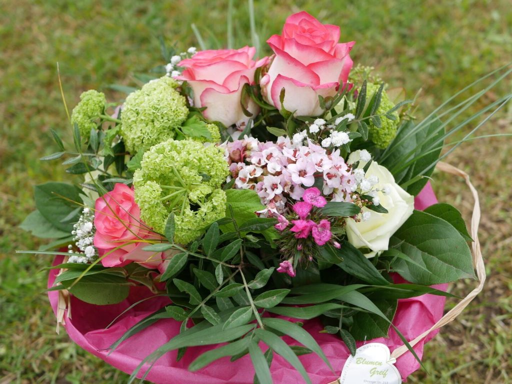 Bunter Blumenstrauß mit rosa Rosen, weißen und rosa Blüten sowie grünen Blättern, auf einem pinken Geschenkpapier, im Freien auf Gras liegend.