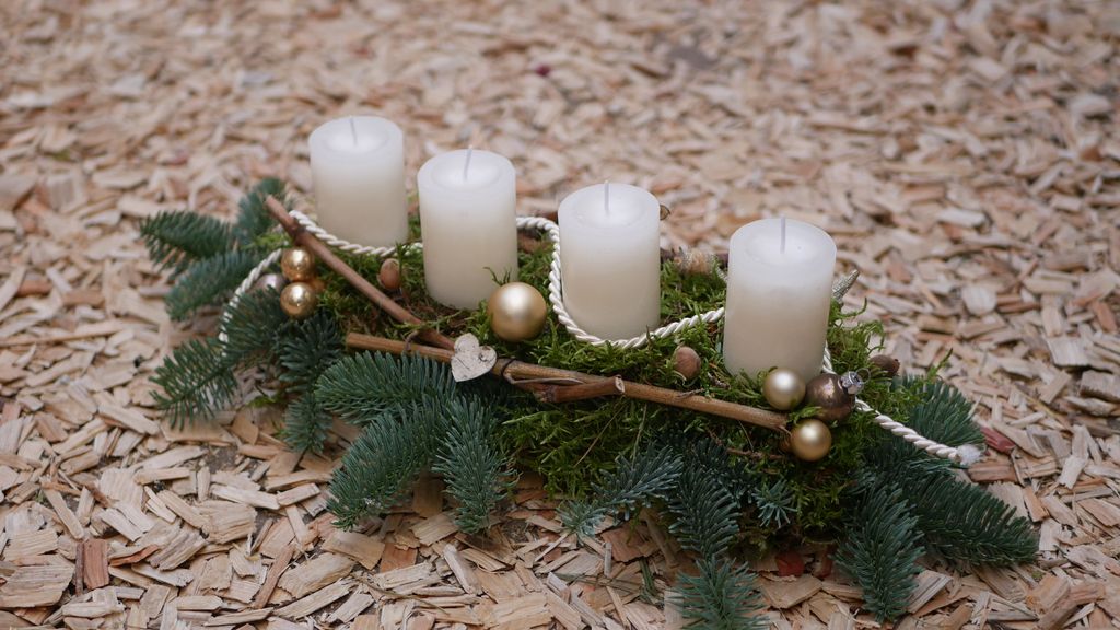 Ein Adventsgesteck mit vier weißen Kerzen, goldenen Kugeln und Tannenzweigen auf Holzspänen.