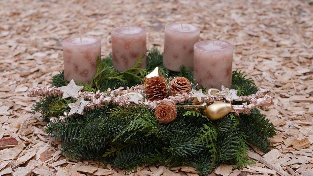 Ein Adventskranz aus Tannenzweigen mit vier rosa Kerzen, verziert mit goldfarbenen und natürlichen Tannenzapfen sowie Sternanhängern, auf gehacktem Holzuntergrund.