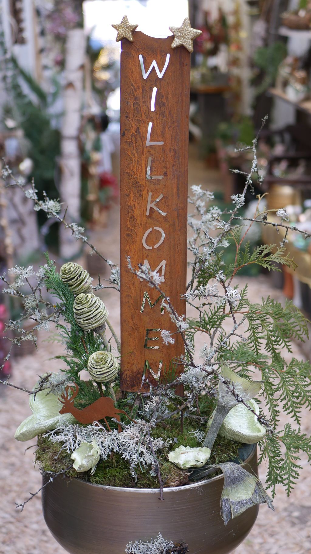 Ein blumiger Willkommensgruß mit einem rostfarbenen Schild, auf dem "WILLKOMMEN" steht, umgeben von grünen und weißen Pflanzen in einem metallischen Übertopf.