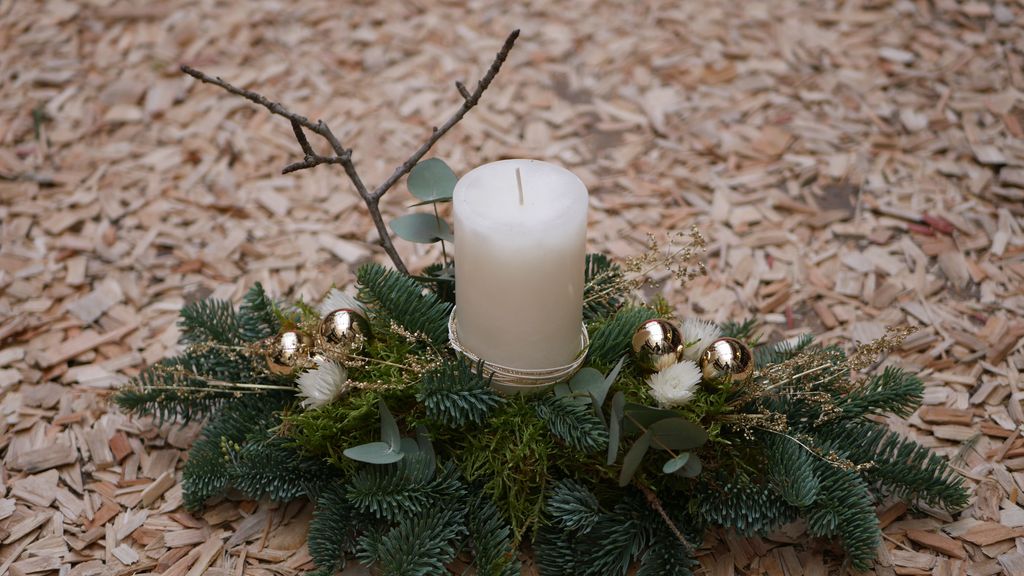 Eine weihnachtliche Dekoration aus Tannenzweigen, goldenen Kugeln, kleinen Zweigen und einer großen weißen Kerze in der Mitte auf Holzhackschnitzeln.