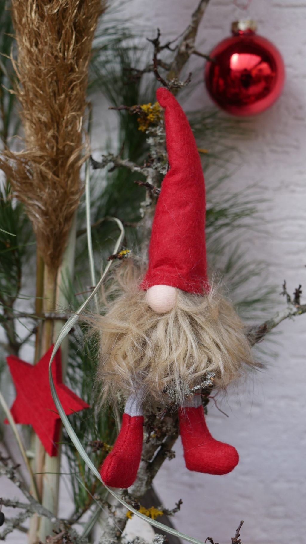 Weihnachtliche Dekoration mit kleinem Wichtel aus rotem Filz, der an einem Ast hängt, im Hintergrund ein roter Weihnachtskugel-Ornament und natürliche Zweige.