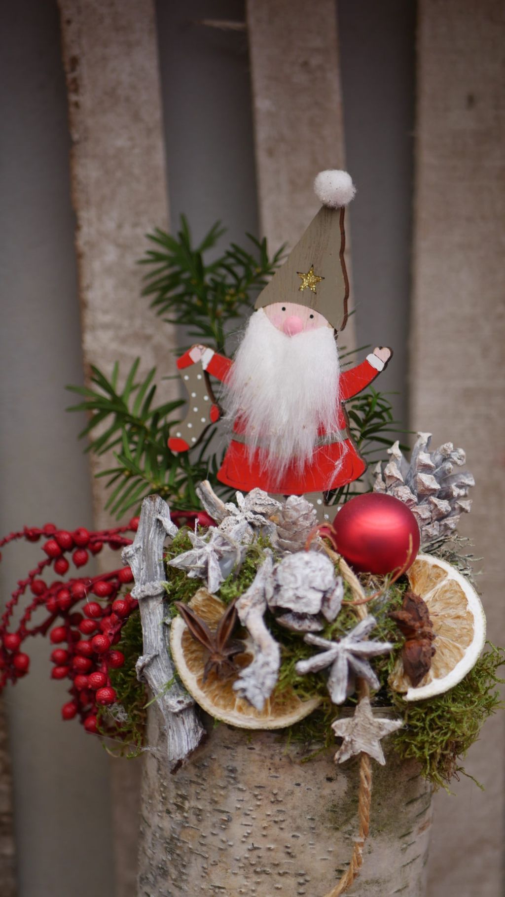 Weihnachtliche Dekoration aus Birkenholz mit einem kleinen Weihnachtsmann aus Holz, Tannenzapfen, roten Beeren, getrockneten Orangenscheiben, Moos und einer roten Weihnachtskugel.