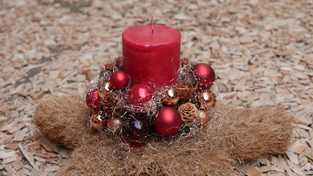 Rote Kerze auf einem dekorativen Kranz mit roten und goldfarbenen Kugeln, kleinen Tannenzapfen und silbernen Schnüren auf einem sternförmigen Stroh- oder Hanfuntergrund auf Holzspänen.