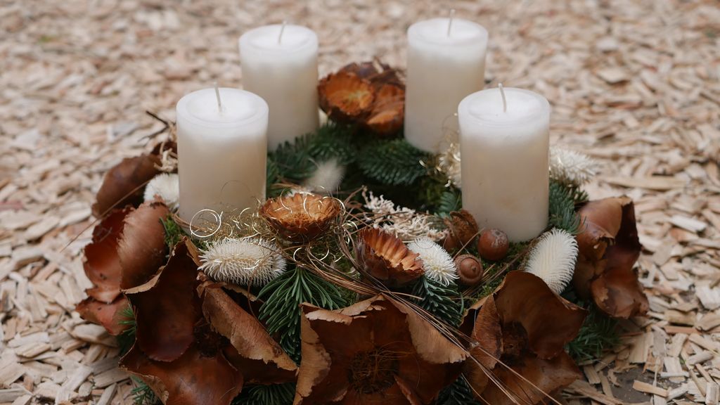 Ein Adventskranz aus Tannenzweigen, dekoriert mit getrockneten Blüten, Zapfen und vier ungezündeten weißen Kerzen, auf einem Boden aus Holzspänen.
