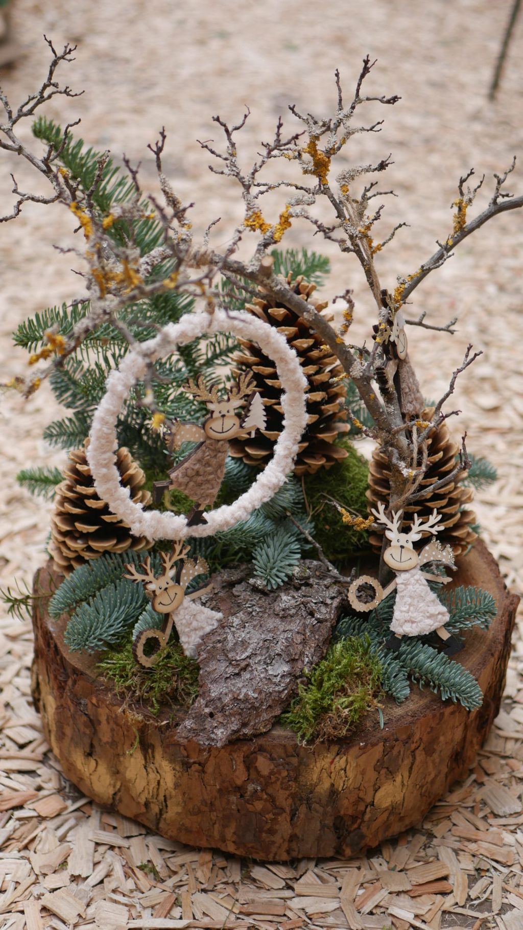 Rustikale Weihnachtsdekoration mit Tannenzapfen, Tannenzweigen, Moos und kleinen handgemachten Rentierfiguren aus Holz und Stoff auf einem Holzstamm.