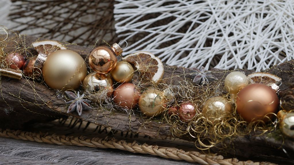 Weihnachtliche Dekoration auf einem braunen Holzstück mit goldenen, kupferfarbenen und perlmuttfarbenen Christbaumkugeln, getrockneten Orangenscheiben, Sternanis und goldfarbenem Deko-Garn.