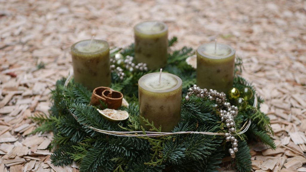 Ein grüner Adventskranz mit vier grünen Kerzen, Zimtstange, getrockneter Orangenscheibe und silbernen Perlen auf Holzspänen gelegt.