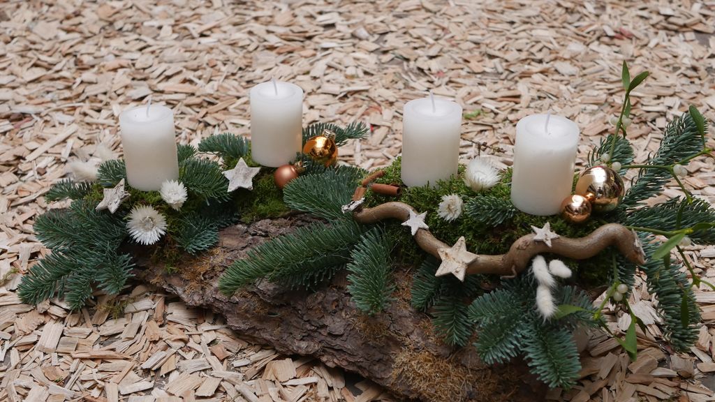 Ein Adventskranz mit vier weißen Kerzen, Tannenzweigen, goldenen Kugeln und weißen Sternen auf einer Baumrinde gelegen, umgeben von Holzspänen.