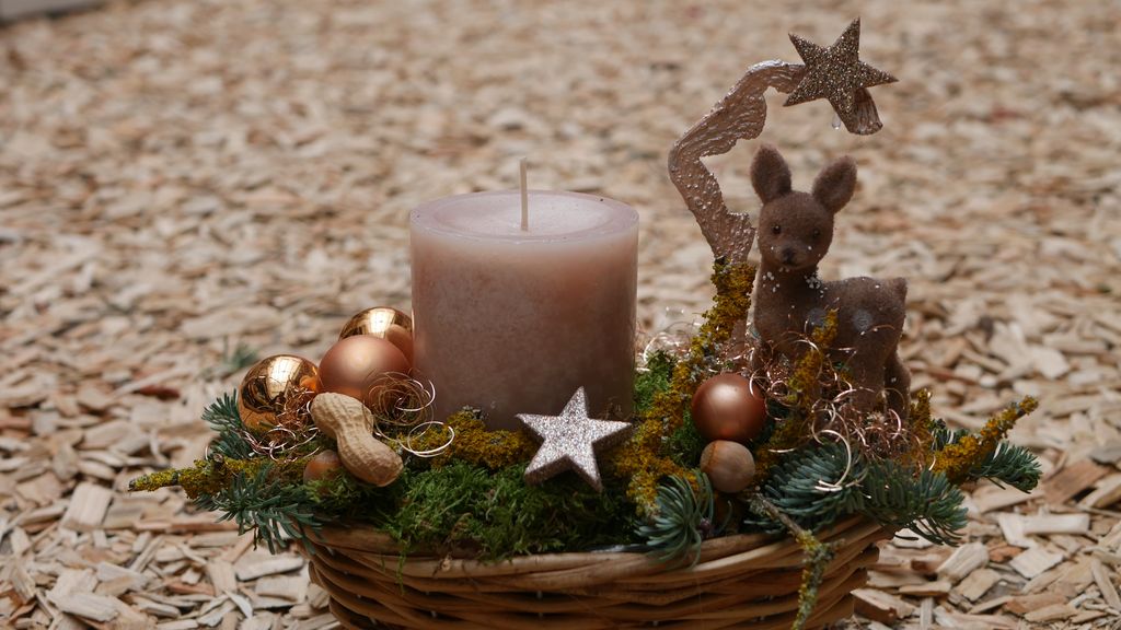 Ein dekorativer Korb mit einer großen braunen Kerze, grünem Moos, goldenen Christbaumkugeln, einer kleinen Dekofigur eines Rehs und einem goldenen Stern auf einem hölzernen Spanplattenboden.