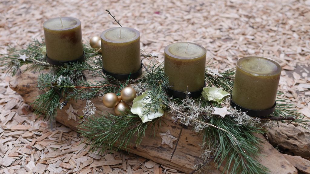 Vier grüne Kerzen auf einem hölzernen Brett, dekoriert mit Tannenzweigen, goldenen Kugeln, weißen Blüten und kleinen Sternverzierungen, liegend auf Holzhackschnitzeln.