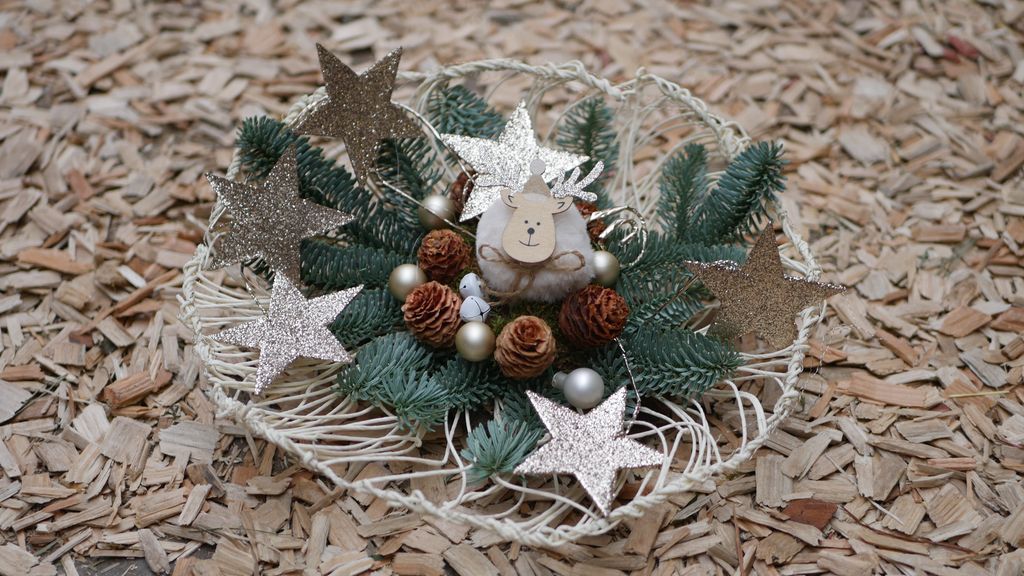 Ein Weihnachtsgesteck mit grünen Tannenzweigen, Tannenzapfen, goldenen Kugeln und glitzernden Sternen, verziert mit einer kleinen Rentierfigur, alles auf einem weißen Drahtkorb vor einem Hintergrund aus Holzspänen.
