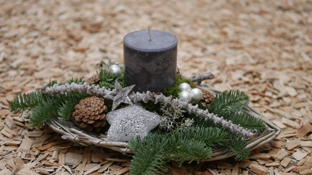 Dekorativer Adventskranz mit schwarzer Kerze, Tannenzweigen, Zapfen und silbernen Sternen auf einem Holzspäneboden.