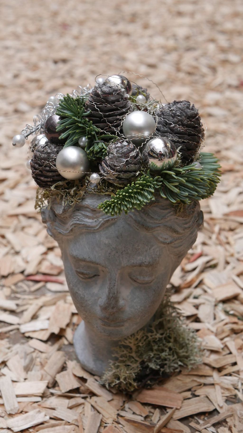 Ein grauer Skulpturkopf mit geschlossenen Augen, der mit Tannenzapfen, silbernen Weihnachtskugeln und Tannenzweigen dekoriert ist, steht auf einem Boden mit Holzhackschnitzeln.