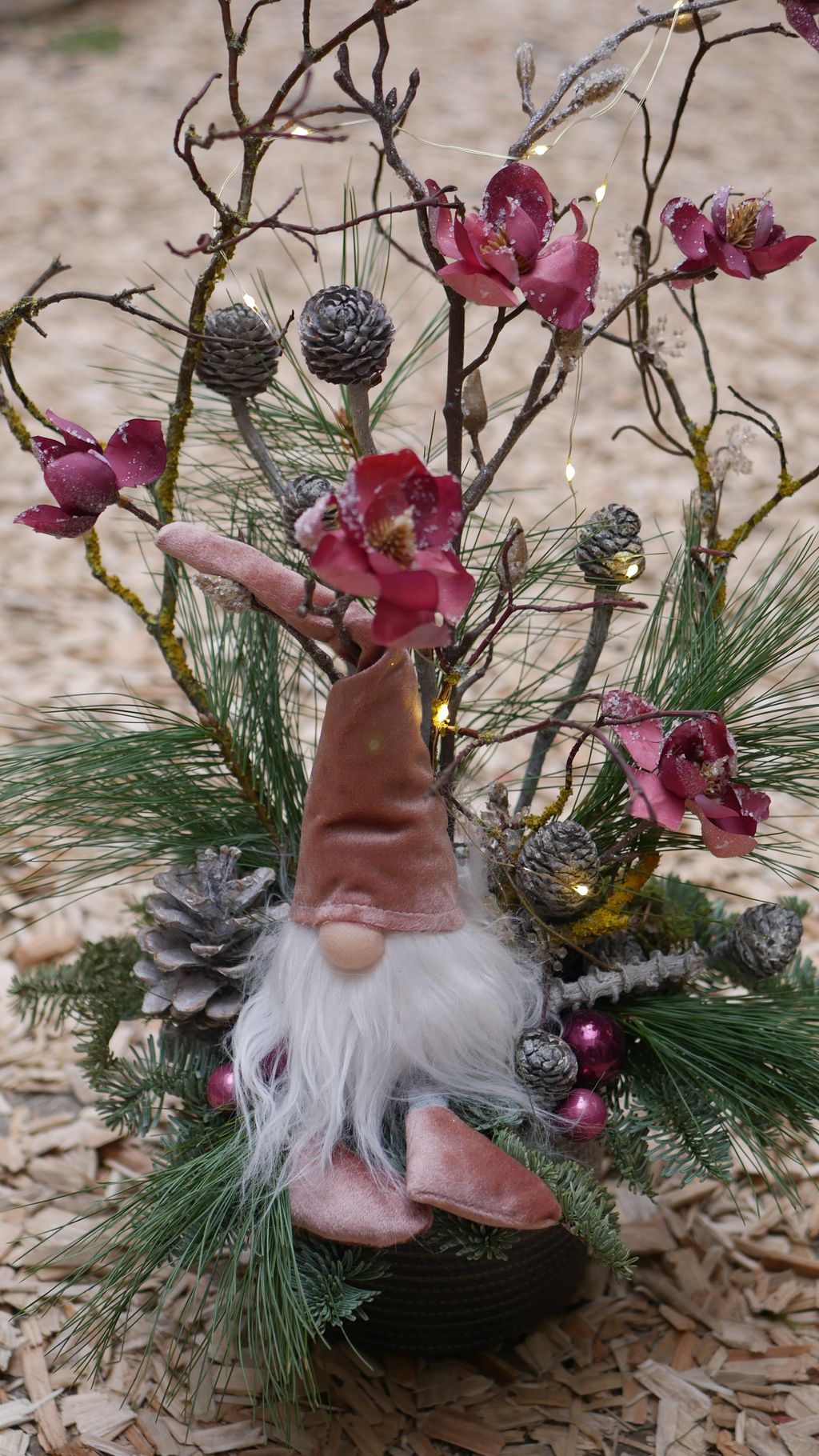 Weihnachtliche Dekoration mit einem Wichtel aus rosa Samt, umgeben von Tannenzweigen, Tannenzapfen, rosa Blumen und kleinen Lichterketten auf Holzspänenboden.