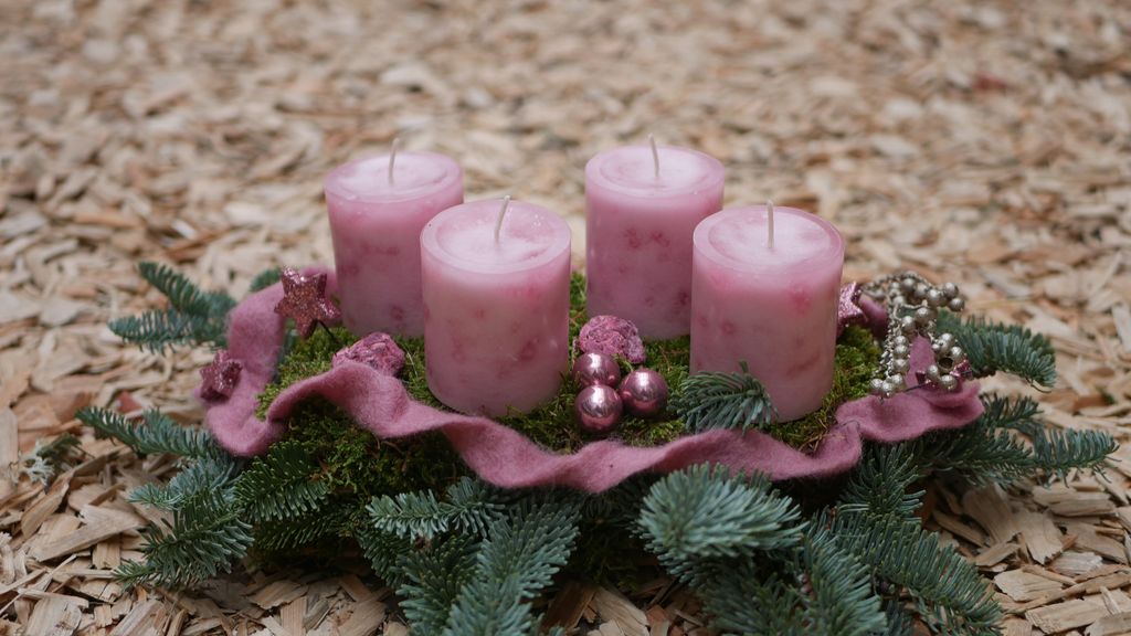 Vier rosa Adventskerzen auf einem grünen Tannengesteck mit Dekorationen wie kleinen Kugeln, Perlen und einem rosa Stoffband auf einem Holzspänteppich.