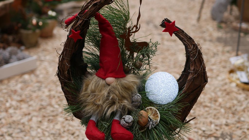 Weihnachtliche Dekoration mit einem kleinen Wichtel in roter Mütze und Stiefeln, umgeben von Tannenzweigen, getrockneten Orangenscheiben und einem glitzernden weißen Weihnachtskugelornament.