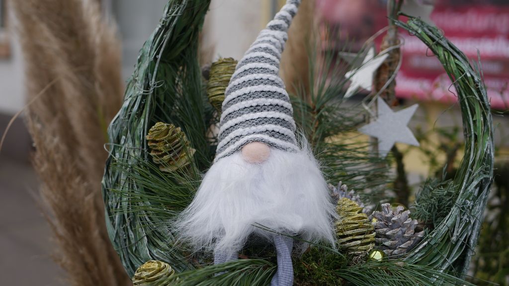 Weihnachtliche Dekoration mit einem kleinen Wichtel mit gestreiftem Mützchen, umgeben von Tannenzweigen und bunt bemalten Zapfen.