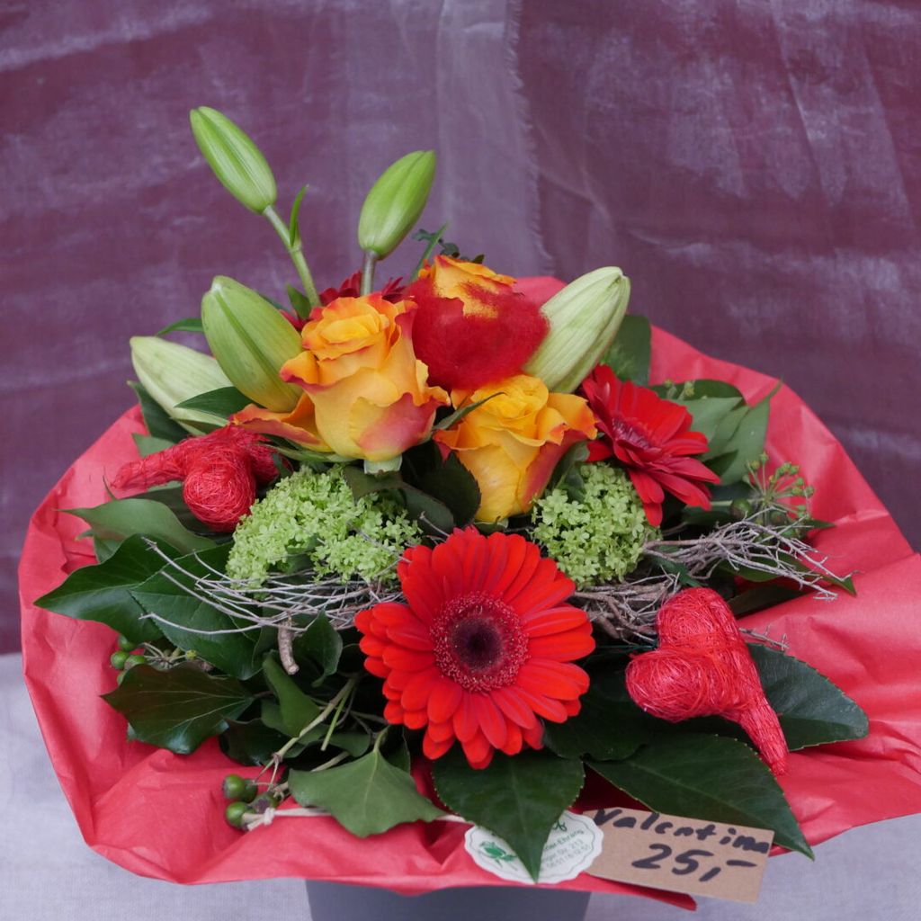 Bunter Blumenstrauß mit gelben Rosen, roten Gerbera, grünen Blättern und Lilienknospen, dekoriert mit roten Herzelementen und auf rotem Papier arrangiert.