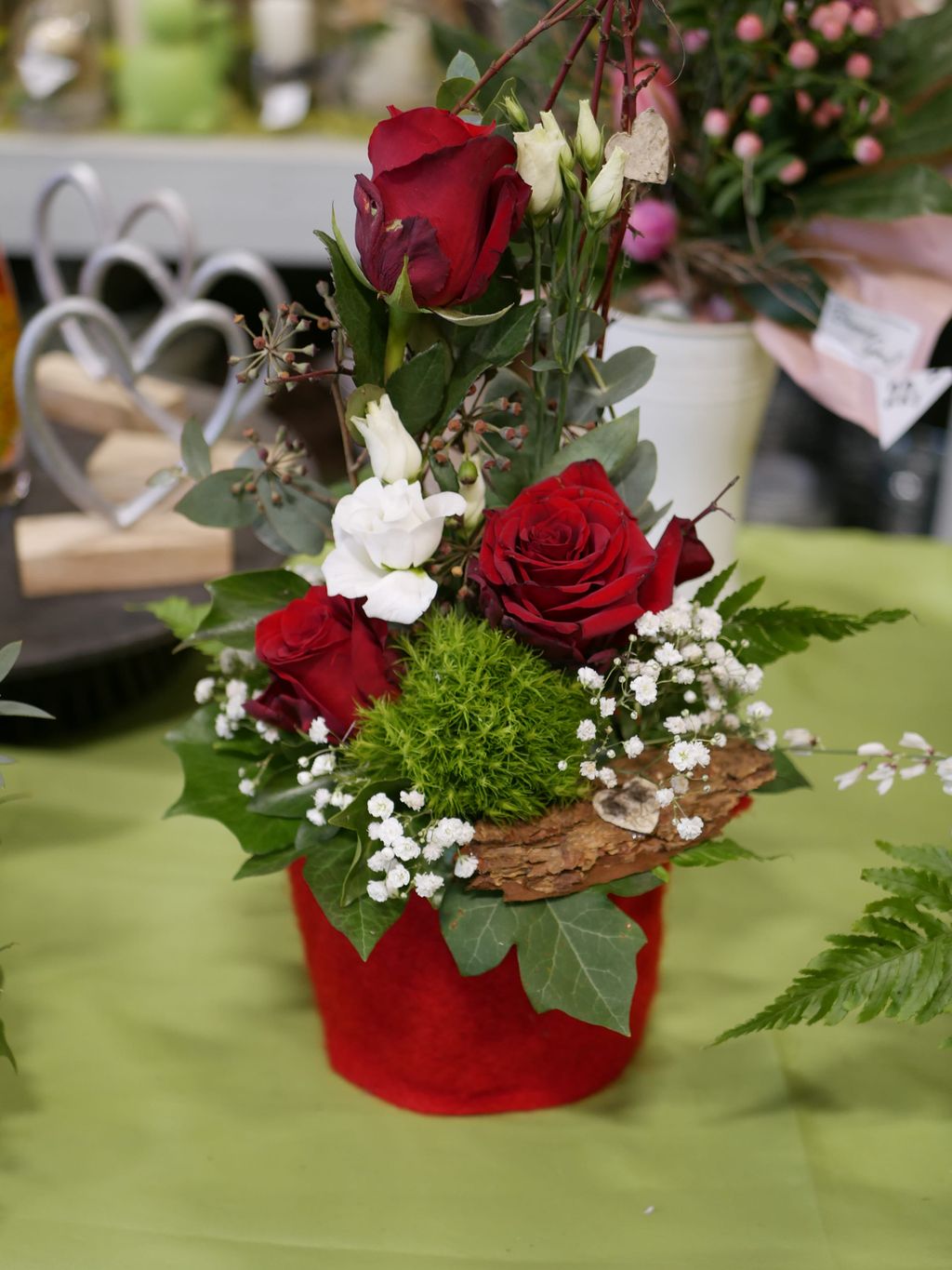 Ein Blumenarrangement in einem roten Übertopf mit roten Rosen, weißen Blüten, grünen Blättern und Dekor aus Rinde und kleinen Herzen aus Holz.