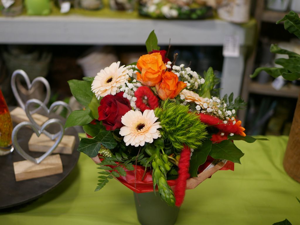 Bunter Blumenstrauß mit orangen Rosen, roten und weißen Blumen sowie grünen Blättern in einem grauen Blumentopf auf einem grünen Tisch.