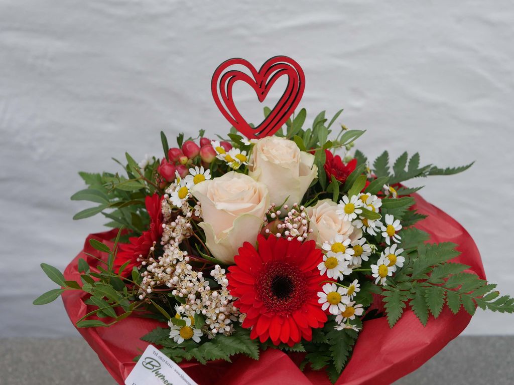 Ein Blumenstrauß mit weißen Rosen, roten Gerbera und kleinen weißen Margariten, dekoriert mit grünen Blättern und einem roten Herzstecker, umgeben von rotem Geschenkpapier.