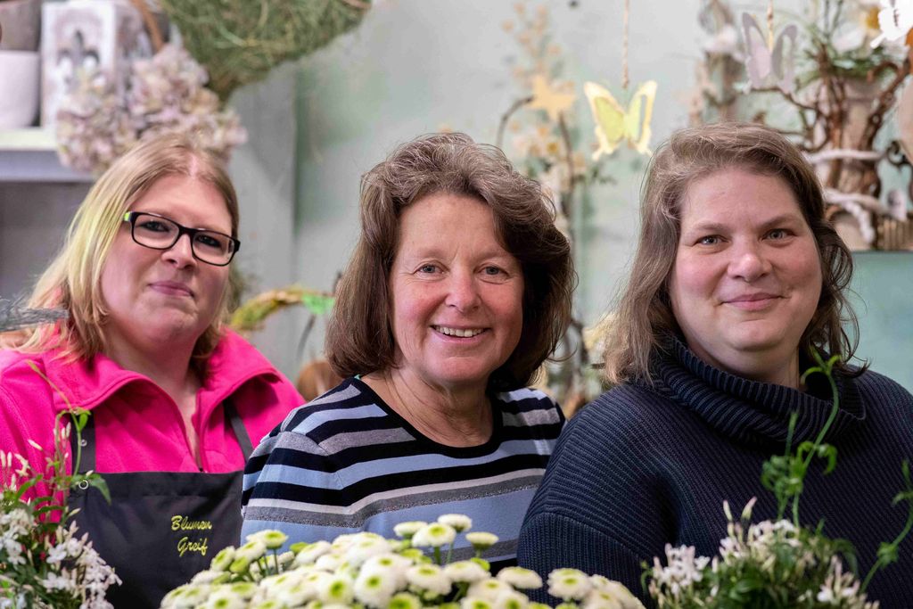 Drei Frauen stehen in einem Blumengeschäft, umgeben von Blumen und Pflanzen, und lächeln in die Kamera.