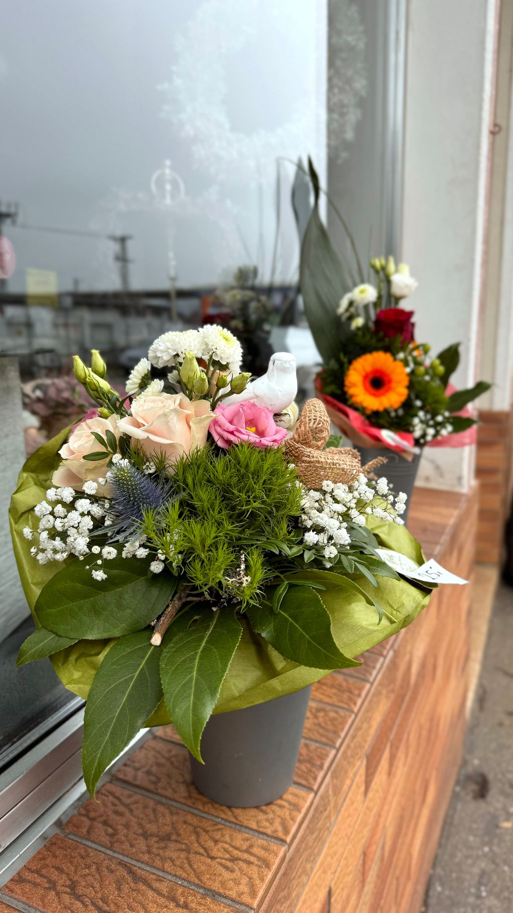 Zwei Blumenarrangements in grauen Vasen auf einer Fensterbank, mit verschiedenen Blumen und grünen Blättern dekoriert.