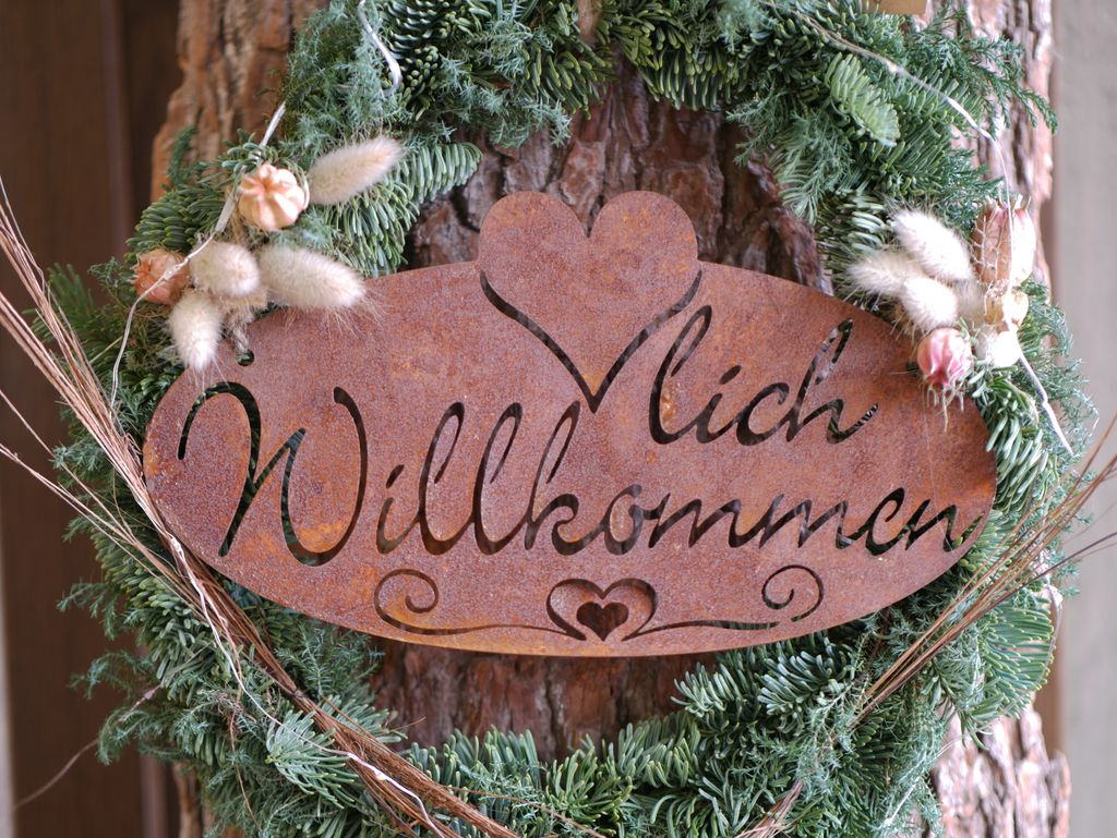 Rostige Metallschild mit der Aufschrift ‚Willkommen‘, umgeben von einem grünen Tannenzweigkranz mit getrockneten Blumen vor einem Baumstamm.