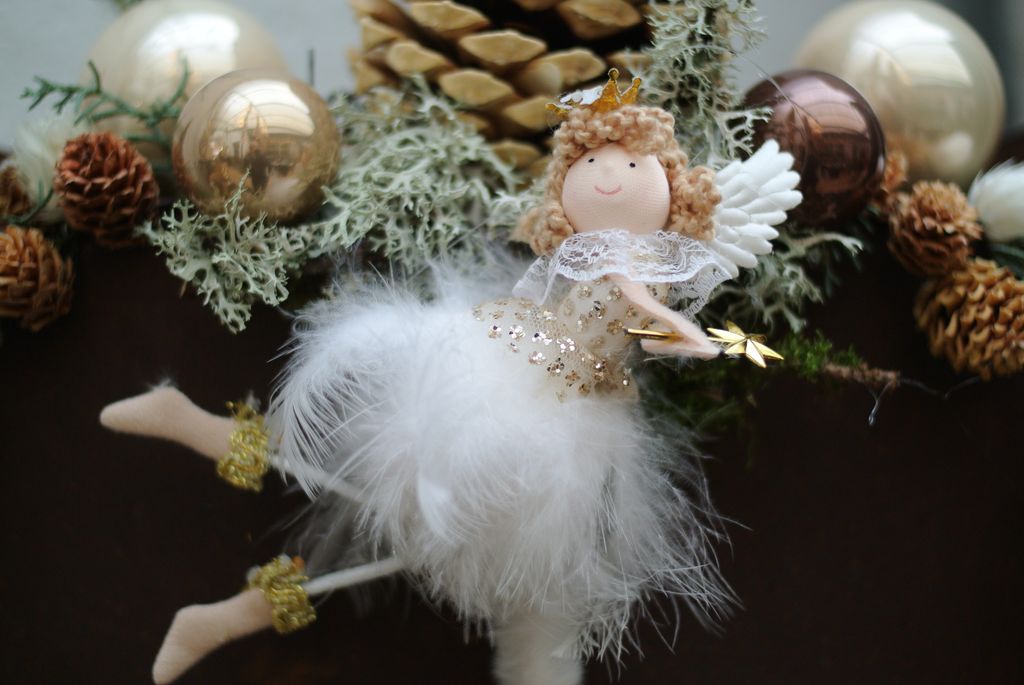 Deko-Engelfigur mit Krone, Flügeln und Federrock vor Tannenzapfen, Moos und goldenen Christbaumkugeln.
