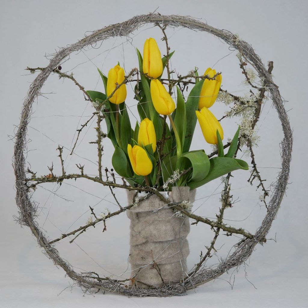 Gelbe Tulpen in einem grauen, miteingewickelten Behälter, umgeben von einem runden Rahmen aus trockenen Zweigen und Spinnweben vor hellem Hintergrund.