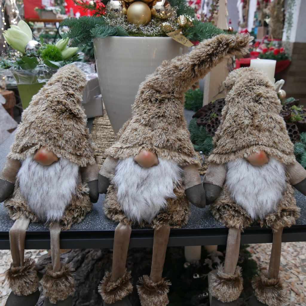 Drei sitzende Wichtel-Figuren mit langen, spitzen und braunen Fellmützen, weißen Bärten und beigen Kleidern auf einem Tisch, im Hintergrund weihnachtliche Dekoration mit Kerzen und Pflanzen.