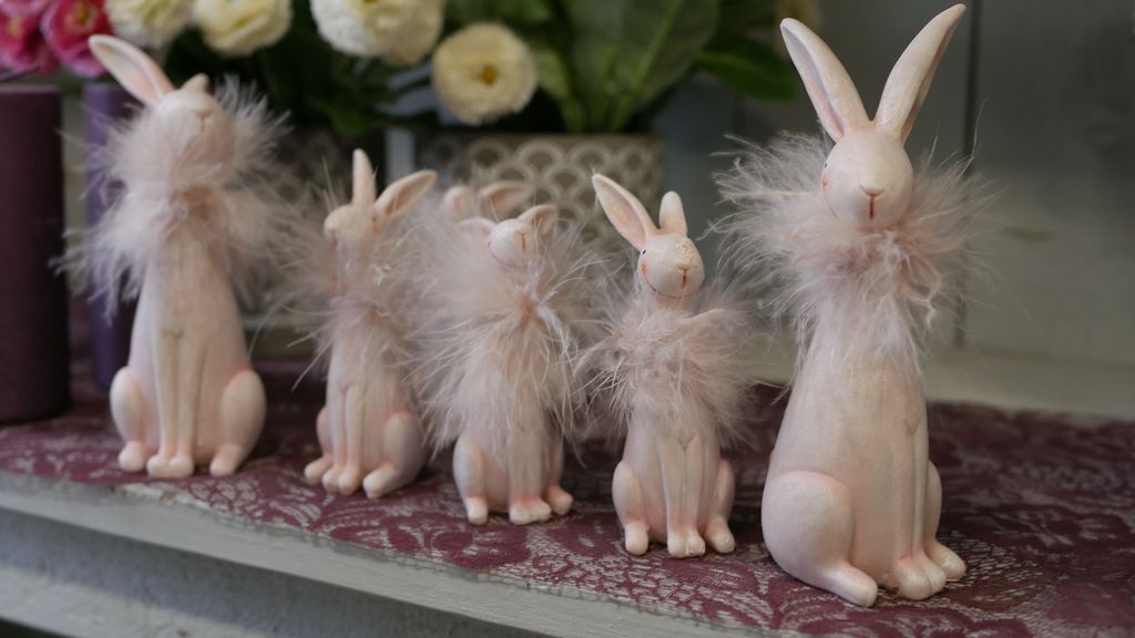 Fünf dekorative Hasenfiguren in verschiedenen Größen mit flauschigen Kragen, die auf einer gemusterten Tischdecke vor einem Blumengesteck stehen.