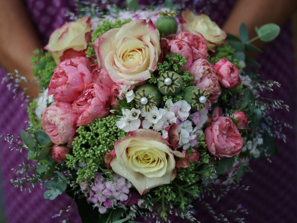 Nahaufnahme eines farbenfrohen Blumenstraußes mit rosa und cremefarbenen Rosen, grünen Pflanzen und kleinen weißen Blüten vor einem violetten Hintergrund.
