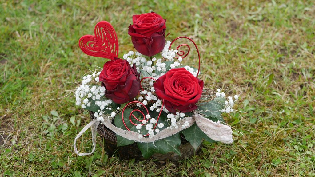 Ein Blumenarrangement mit drei roten Rosen, weißen Gipskraut-Blüten und grünen Blättern auf Grasboden, verziert mit roten Herz-Dekorationen und kleinen Spiralen aus Draht.