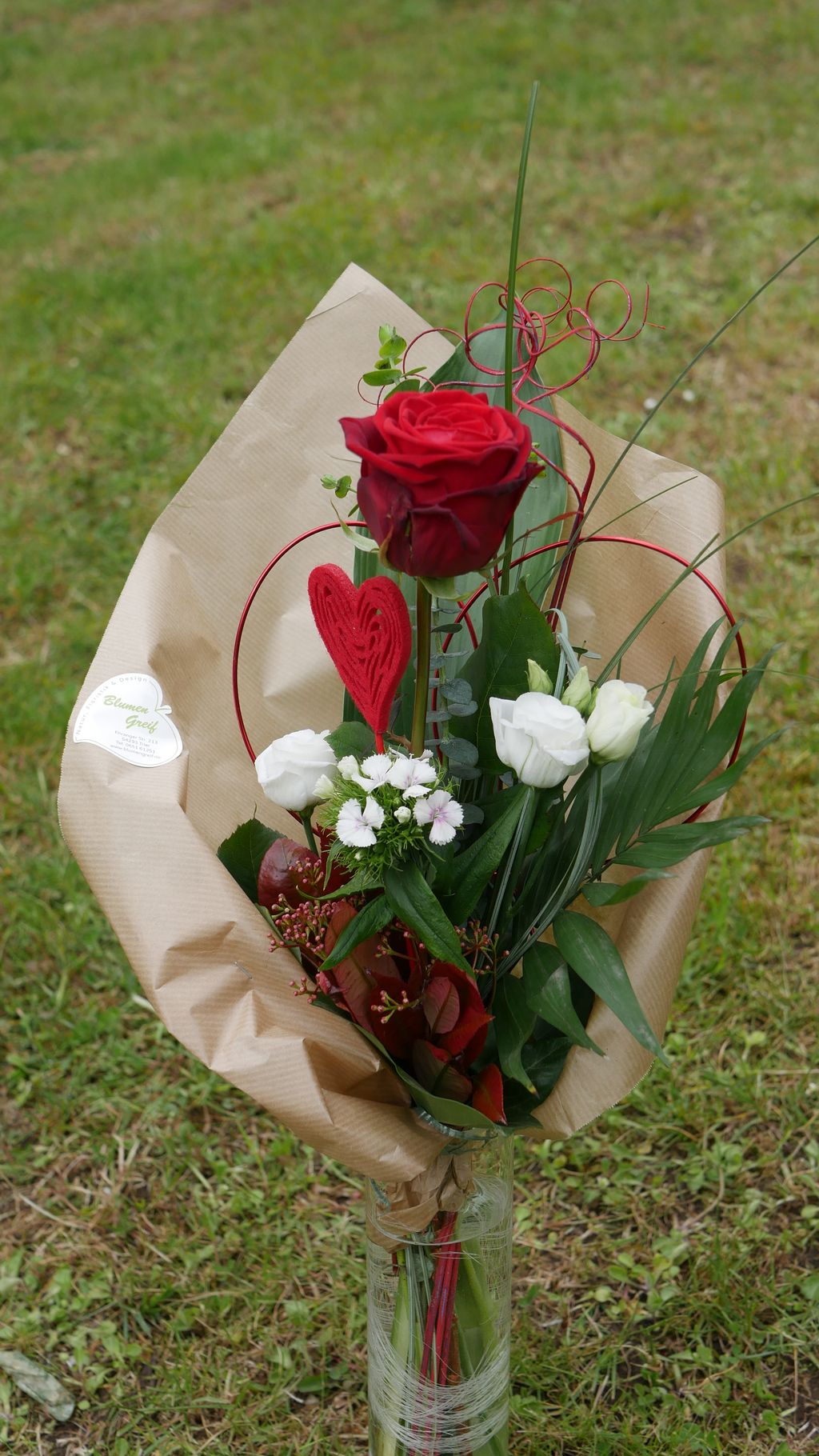 Ein Blumenstrauß mit einer roten Rose, weißen Blüten, grünen Blättern und einem roten Herz aus Draht, umhüllt von braunem Papier, in einer Glasvase auf einer Wiese.