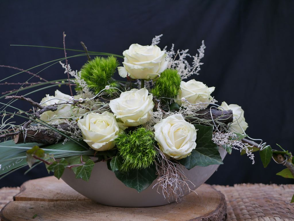 Ein Blumenarrangement mit weißen Rosen, grünen Kugelpflanzen und verschiedenen Zweigen in einer weißen Schale, platziert auf einem Baumstamm vor dunklem Hintergrund.
