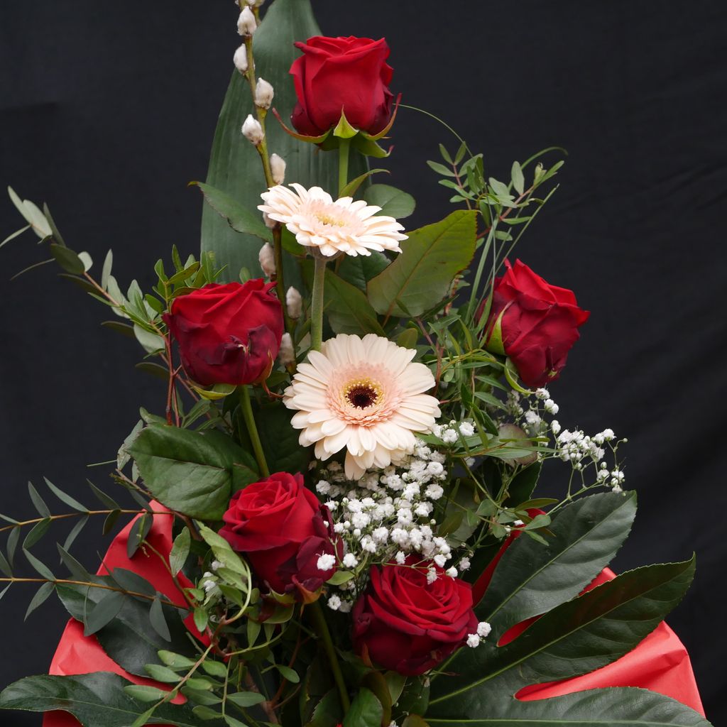 Strauß mit fünf roten Rosen, zwei weißen Gerbera und weiterem Grün sowie kleinen weißen Blüten, vor schwarzem Hintergrund und rotem Stoff dekoriert.