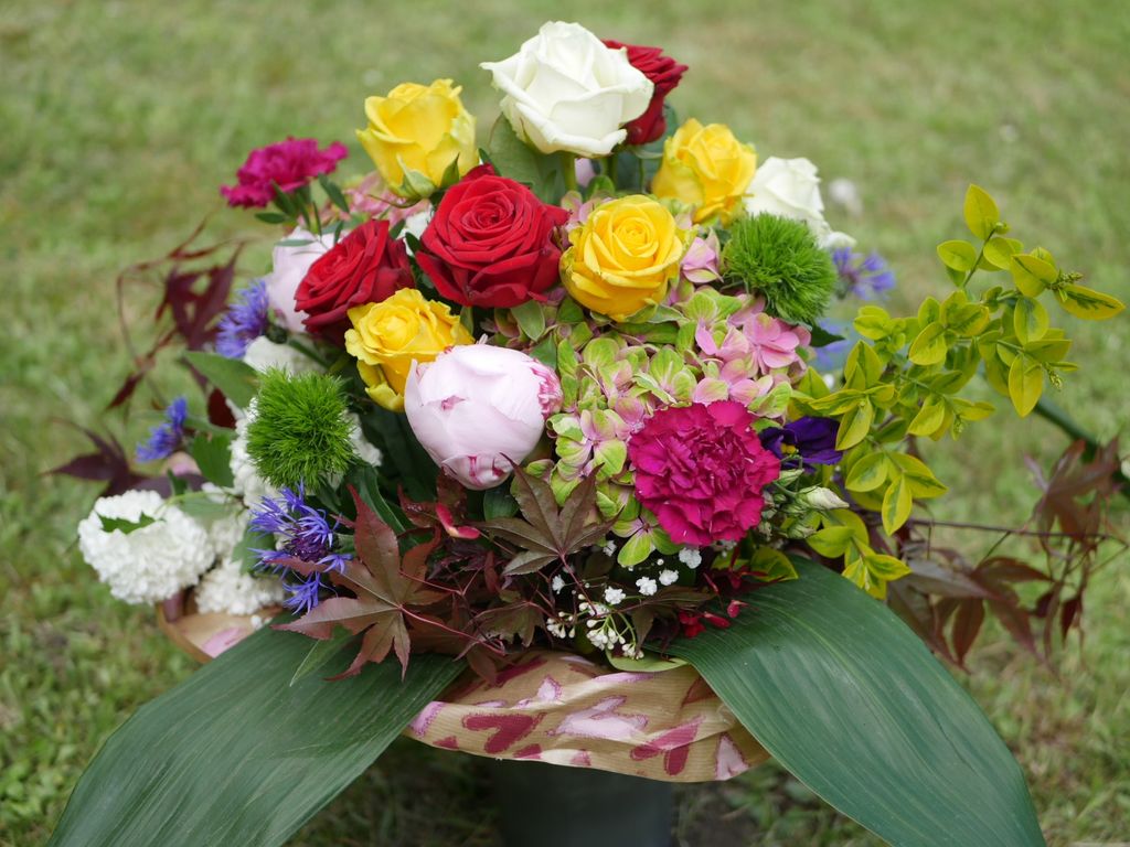 Bunter Blumenstrauß mit roten, gelben, weißen und pinkfarbenen Rosen sowie verschiedenen grünen Blättern und anderen Blumen auf einem grünen Grasuntergrund.